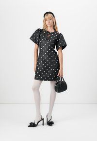 Vrouw in een zwart jurk met stippen en fluttermouwen, witte tights, zwart strikhakken, een haarband en een zwart gewatteerd handtasje.