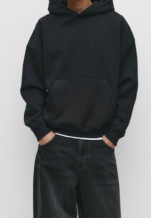 Person iført en sort hættetrøje med forlommer og sorte jeans, hænderne i lommerne, stående mod en ensfarvet lys baggrund.
