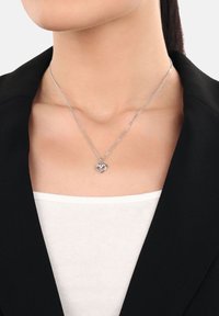 Donna che indossa una collana con ciondolo a forma di cuore in argento, con una piccola pietra trasparente, abbinata a un blazer nero e a una maglietta bianca.