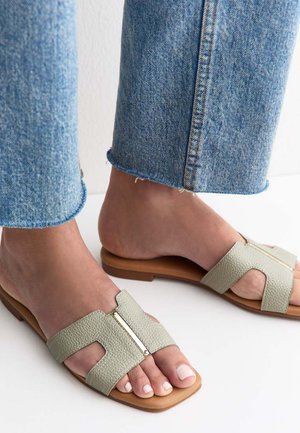 Pies con sandalias tipo slide de color verde claro texturizadas con detalle dorado en suelas tostadas, combinadas con vaqueros azules recortados sobre suelo blanco.