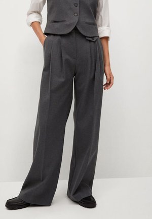 Frau trägt hoch taillierte, weit geschnittene, dunkelgraue Hose, weißes Hemd mit hochgekrempelten Ärmeln, dunkelgraue Weste und schwarze Schuhe, steht vor weißem Hintergrund.