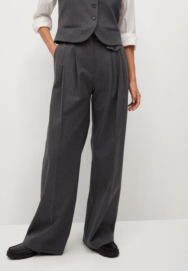 Femme portant un pantalon large taille haute gris foncé, une chemise blanche aux manches retroussées, un gilet gris foncé et des chaussures noires, debout devant un fond blanc.