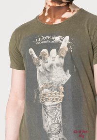 T-shirt vert olive avec un imprimé graphique d'une main ornée de bijoux, de chaînes et du texte "Laissez vos soucis derrière vous" en noir décoloré.