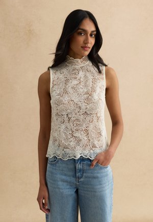 Frau mit langen dunklen Haaren, die ein ärmelloses weißes Spitzenoberteil mit Blumenmuster und blaue Jeans trägt, stehend vor einem beigen Hintergrund.