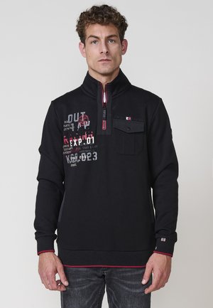 Sudadera negra con cuello alto, cremallera destacada y acentos rojos. Texto gráfico en la parte delantera y un bolsillo de parche en el pecho. Puños y dobladillo acanalados.