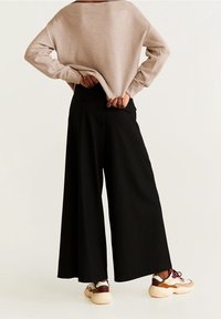 Übergroßer beiger Pullover, weite schwarze Hose und klobige Sneakers mit kastanienbraunen Akzenten. Weicher Stoff und lockere Passform mit sichtbaren Gesäßtaschen.