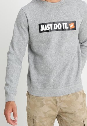 Mężczyzna noszący szarą bluzę z czarno-białym napisem "JUST DO IT." i pomarańczowym logo Nike, zestawioną z beżowymi bojówkami w kamuflażu.