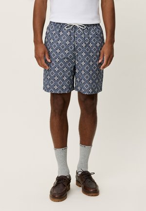 Tumesinised ujumispüksid, millel on keerukas valge lille- ja teemantmuster. Omavad nööriga vöökohaga ja kerges kangas. Kantud hallide sokkide ja pruunide kingadega.