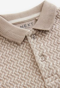 Camisa polo texturizada de color beige con cuello de punto, que presenta un patrón geométrico y dos botones marrones en el cuello.