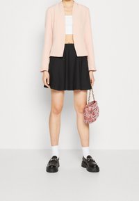 Žena v růžovém saku, bílém crop topu, černé sukni, bílých ponožkách a černých mokasínech, držící svým bokem růžovou kabelku na řetízku.