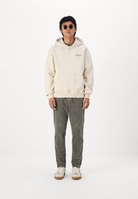Beige Hoodie mit einer Fronttasche und Logo, kombiniert mit grauen Cordhosen. Die Accessoires umfassen dunkle Sonnenbrillen und weiße Sneakers mit braunen Sohlen.