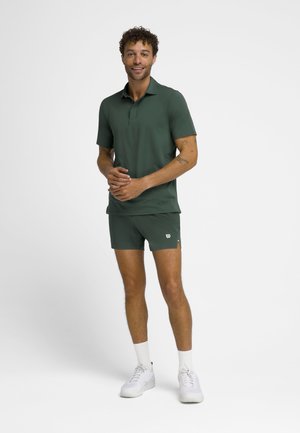 Mand stående iført mørkegrøn kortærmet polo, matchende shorts, hvide sokker og hvide træningssko mod en hvid baggrund.