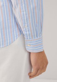 Camisa de rayas azul claro con rayas blancas, naranjas y marino. El puño tiene dos botones y un patrón de rayas complementario.