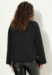 Maglietta nera a maniche lunghe con orlo leggermente curvato e polsini arrotolati. Presenta una texture liscia e un design semplice e lineare.