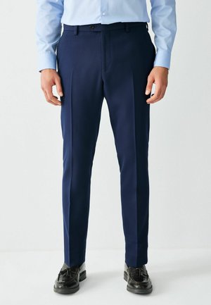 Mężczyzna ubrany w dopasowane granatowe eleganckie spodnie, czarne mokasyny z chwostami oraz jasnoniebieską koszulę na guziki, stoi prosto.