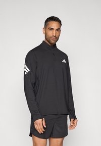 Sort, langærmet atletisk top med lynlåslukning ved kraven og hvid Adidas-logo; har diskrete syninger og tætsiddende design. Kombineret med sorte shorts.