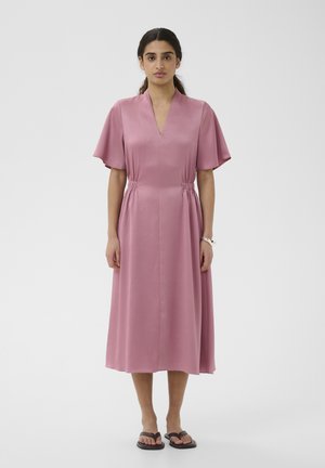 Naine kannab lahtist, keskmise pikkusega tolmroosat kleiti, millel on lühikesed, laiad varrukad ja V-kaelus, ning mustade sandaalide ja valge käevõruga.