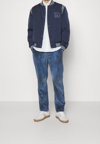 Man i marinblå varsityjacka, vit skjorta, blå kordbyxor och vita sneakers som står med händerna i fickorna.