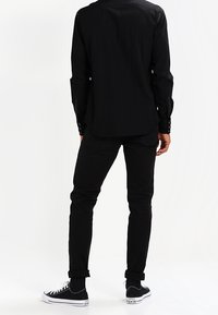 Camicia nera a maniche lunghe con polsini a bottone, abbinata a pantaloni neri slim-fit e sneaker nere basse con dettagli bianchi sulla suola, vista da dietro.