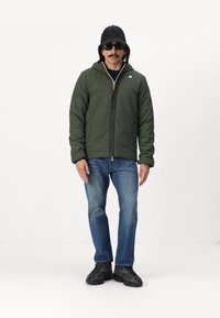 Grøn quiltet jakke med lynlås og hætte, båret over en mørk skjorte; parret med blå jeans og sorte sneakers. Personen bærer solbriller og en sort kasket.
