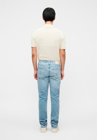 Ανοιχτό μπεζ κοντομάνικο μπλουζάκι σε συνδυασμό με ανοιχτό μπλε τζιν παντελόνι. Το τζιν έχει ραμμένες λεπτομέρειες και ένα τετράγωνο ταμπελάκι στο πίσω μέρος.