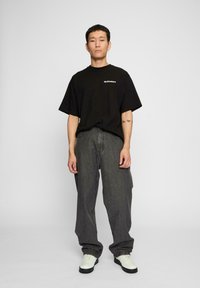 Svart oversized t-skjorte med hvitt logo, sammen med løse grå jeans. Modellen har på seg hvite sneakers med svarte såler. Enkel bakgrunn.