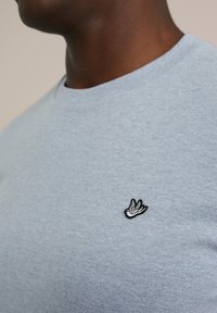 Tricou din bumbac de culoare albastru deschis, cu guler rotund și un mic logo brodat în formă de pasăre neagră pe partea stângă a pieptului. Textură netedă.