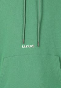 Mintgrüner Hoodie mit einer Vordertasche, ausgestattet mit Kordeln und silbernen Metallenden. "LES'ARCS" ist in Weiß über der Tasche aufgedruckt.