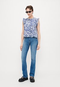 White top with blue floral pattern, ruffled shoulder seams, paired with light denim flared jeans and black chunky platform shoes.
