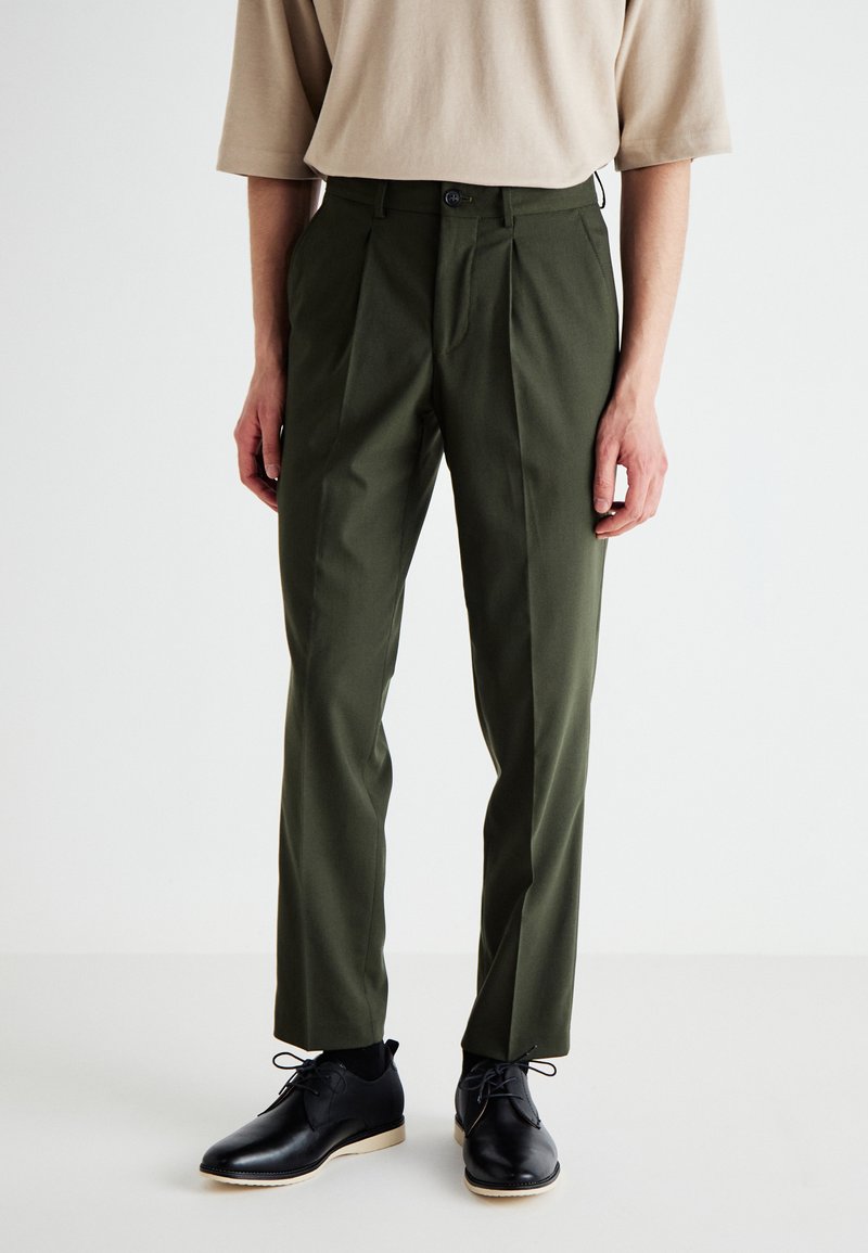 Pantalons plissés verts en tissu lisse, présentant une coupe slim et une fermeture à bouton à l'avant, associés à des chaussures de ville noires.