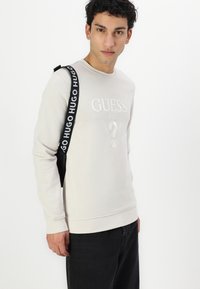 Sudadera de color beige claro con puños y dobladillo acanalados, que presenta un logo blanco en relieve y un gráfico de signo de interrogación. Combinada con jeans oscuros.