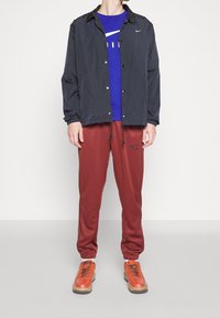 Black lightweight jacket with snaps, blue Nike sweatshirt, maroon joggers with side stripes, and orange sneakers with brown soles.