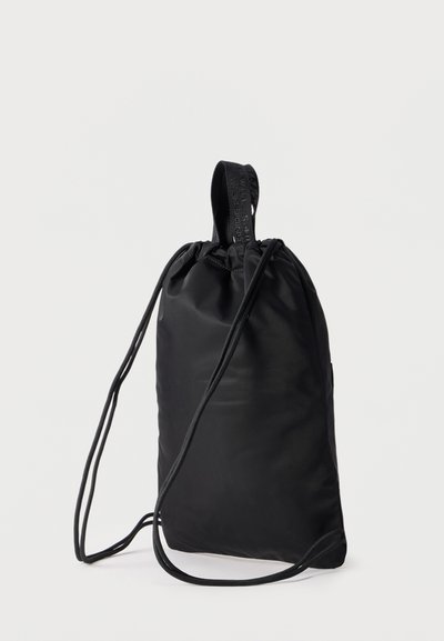 Μαύρο σακίδιο με κορδόνι, υφασμάτινη λαβή και λεπτά ρυθμιζόμενα λουριά ώμου, όρθιο σε λευκό φόντο.