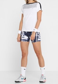 Valkoinen urheilut-paita, jossa harmaita ja laivastonsinisiä yksityiskohtia, yhdistetty laivastonsinisiin shortsseihin, joissa on vaalea kuvio. Mukana valkoiset lyhyet sukat ja lenkkarit.