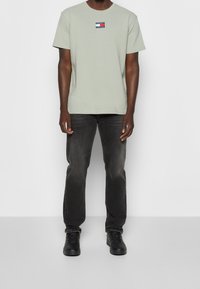 Camiseta de algodón verde claro con un pequeño logo de marca en rojo, blanco y azul en el pecho, combinada con unos vaqueros gris oscuro y zapatillas negras.