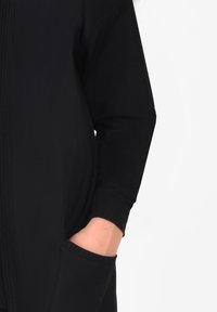 Svart långärmad plagg med ribbade muddar och en framficka. Slät struktur och avslappnad passform, med fina vertikala sömndetaljer.