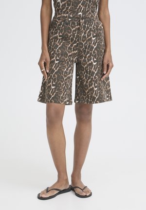 Person iført knælange shorts med leopardprint og sorte klipklappere, stående med hænderne afslappet ned langs siderne.