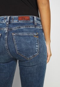 Jeans de mezclilla azul oscuro con un diseño de tiro medio, costuras visibles, dos bolsillos traseros y una etiqueta de cuero marrón con "LTB" en la cinturilla.