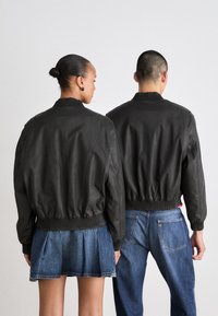 Sorte bomberjakker med ribbede kraver og manchetter, båret af to modeller. Den ene er parret med en denimnederdel, mens den anden har blå jeans på.