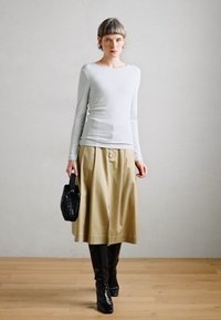 Top de mangas longas cinza claro, saia plisada bege, botas pretas até ao joelho e uma mala preta texturizada. Materiais suaves e estruturados.