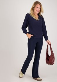 Vrouw in een marineblauwe top met lange mouwen en broek, lopend en glimlachend, met een bordeauxrode handtas met zilveren oogjes, en crèmekleurige sneakers met bordeauxrode strepen.