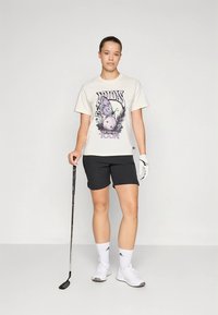 Balti marškinėliai su spalvingu drugelio paveikslėliu, juodi šortai, balti kojinės ir batai. Kairie ranka laikomas golfo klubas, dėvima golfo pirštinė.