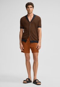 Maglietta a maniche corte a righe marrone e arancione, con colletto nero, abbinata a pantaloni corti color ruggine e infradito nere.