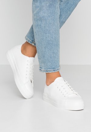 Person iført lyseblå jeans og hvide lave platform sneakers står på en grå gulv mod en ensfarvet baggrund.