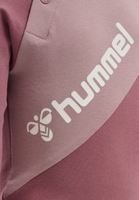 Hummel sweatshirt i en gradient av ljus och mörk rosa, med framträdande vit logotyp och bi-grafik. Slät, texturerad tyg med knappar.