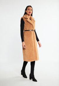 Helle, braune Kunstfellweste mit Schalkragen, am Taillenbereich mit einem schwarzen Lederband gebunden. Getragen über einem schwarzen Langarmoberteil und Leggings.