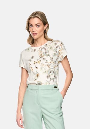 Woman with blonde hair wearing a white floral short-sleeve top and light green high-waist trousers with her hand in pocket.