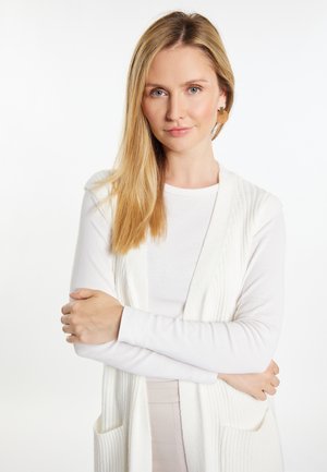 Valkoinen ridullinen neulecardigan valkoisen pitkähihaisen paidan päällä, jossa on huivikaulus ja kaksi etutaskua. Silkkinen tuntuma, vaaleat värit.
