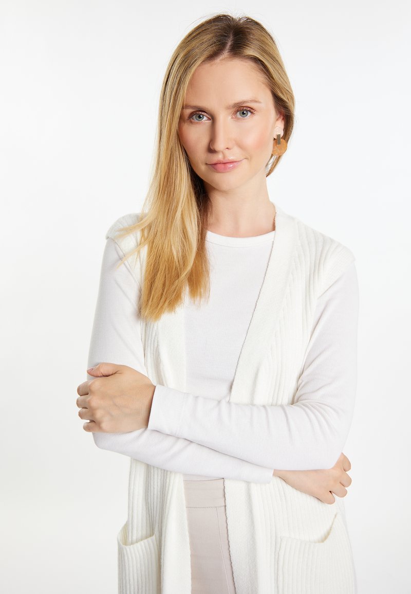 Cardigan alb cu fante ribbed, purtat peste un top alb cu mânecă lungă, având un guler de tip șal și două buzunare frontale. Textură netedă, culoare deschisă.