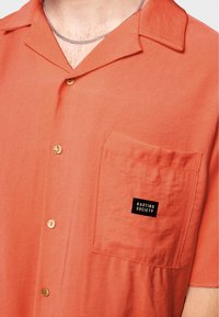 Kortärmad skjorta i korall, tillverkad av mjukt tyg. Har en bröstficka med en svart etikett som står "KAOTIKO SOCIETY" och knappstängning.